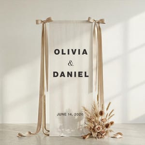 May include: A wedding welcome sign featuring the names "OLIVIA & DANIEL" in black text on a cream-colored fabric. The sign is suspended from a gold metal frame with beige ribbons and a floral arrangement at the base. The date "JUNE 14, 2026" is also displayed.