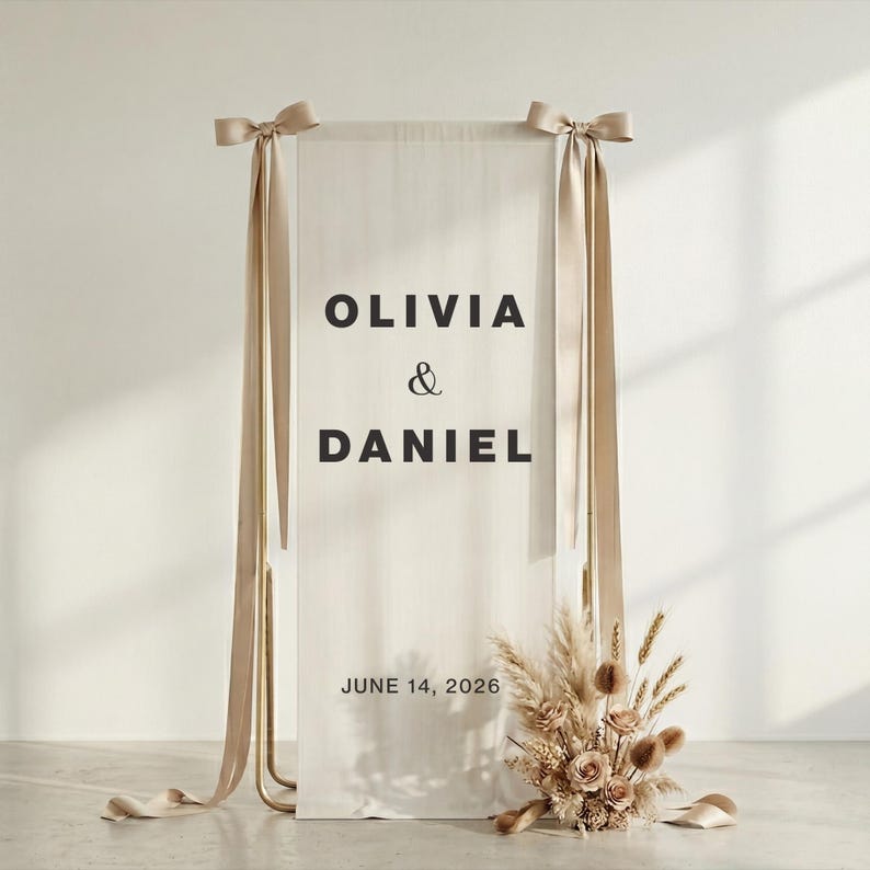 May include: A wedding welcome sign featuring the names "OLIVIA & DANIEL" in black text on a cream-colored fabric. The sign is suspended from a gold metal frame with beige ribbons and a floral arrangement at the base. The date "JUNE 14, 2026" is also displayed.