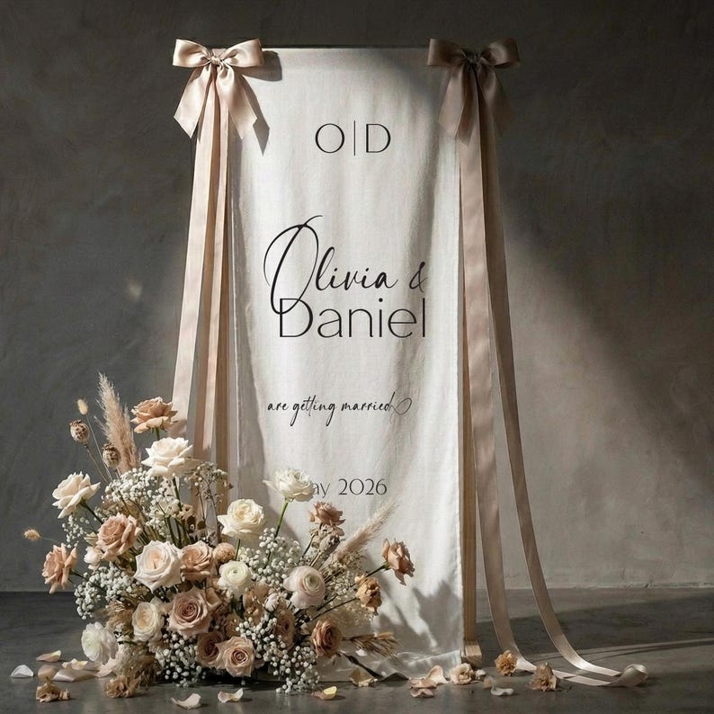 May include: A white wedding welcome sign with the names "Olivia & Daniel" in elegant script, accented by peach-colored ribbons and a floral arrangement of roses and dried flowers. The sign also includes the date "May 2026".