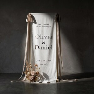 May include: A white fabric wedding welcome sign with the text "WELCOME to the wedding of Olivia & Daniel JUNE 14, 2026 New York". The sign is decorated with beige ribbons and a floral arrangement.