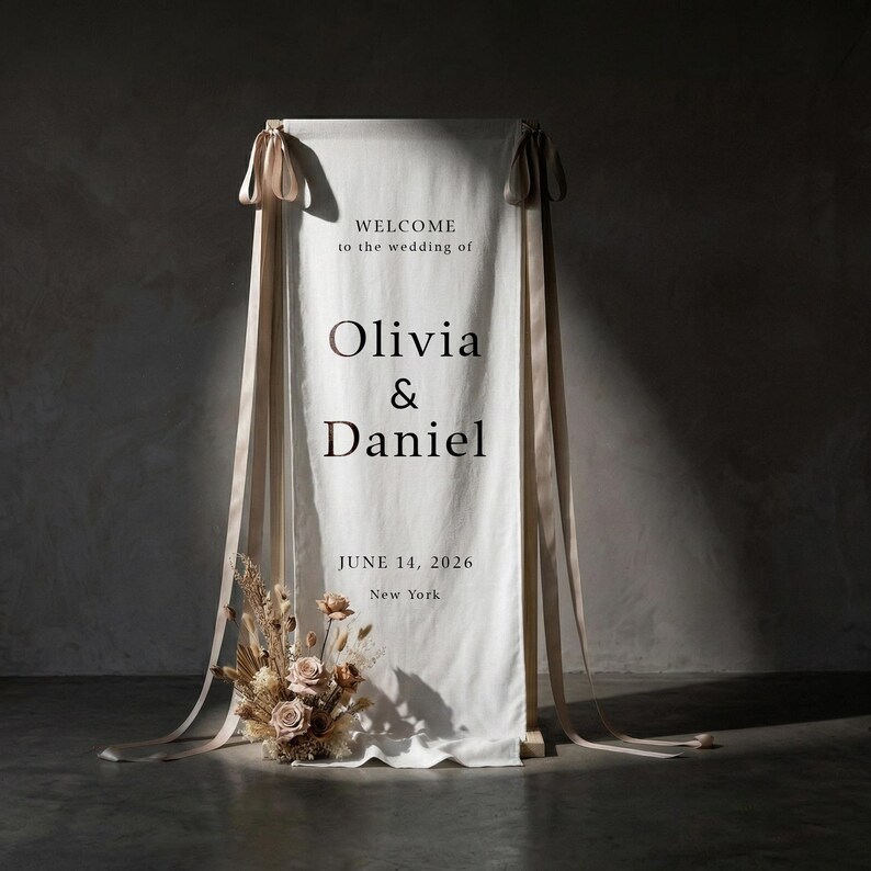 May include: A white fabric wedding welcome sign with the text "WELCOME to the wedding of Olivia & Daniel JUNE 14, 2026 New York". The sign is decorated with beige ribbons and a floral arrangement.