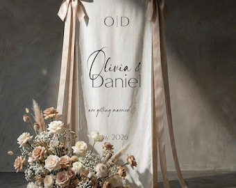 Może przedstawiać: Biały znak powitalny na wesele z imionami "Olivia & Daniel" w eleganckim skrypcie, zaakcentowany aranżacją kwiatową z róż i suszonych kwiatów. Znak ozdobiony jest beżowymi wstążkami i kokardami, z datą "Maj 2026" na dole.
