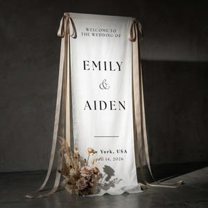 May include: A white wedding welcome sign with the text "WELCOME TO THE WEDDING OF EMILY & AIDEN". The sign is decorated with beige ribbons and a floral arrangement at the base. The date is April 14, 2026.