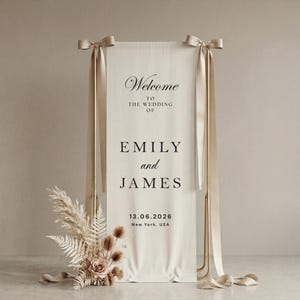 May include: A wedding welcome sign with the names Emily and James, and the date 06/13/2026. The sign is cream-colored with gold accents and ribbons. Dried flowers and pampas grass are at the base of the sign.