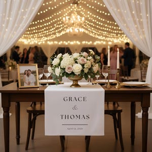 May include: A wedding reception table is set with a floral arrangement, candles, and a table runner. The table runner reads "GRACE & THOMAS" with the date "April 14, 2026". A framed photo of a couple is on the table.