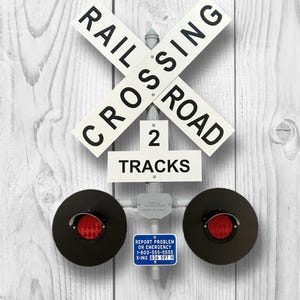 May include: A railroad crossing sign with the words "RAILROAD CROSSING" in black on white crossbars. Below, a sign reads "2 TRACKS." Two black circular lights with red reflectors are present. A blue sign reads "REPORT PROBLEM OR EMERGENCY 1-800-555-5555 X-ING 836 597 H."