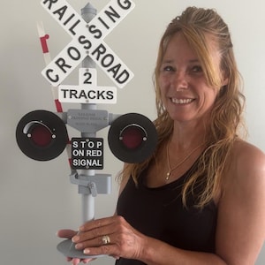 May include: A gray and white railroad crossing sign with the words "RAILROAD CROSSING" and "2 TRACKS". The sign has two circular red lights and a sign that says "STOP ON RED SIGNAL".
