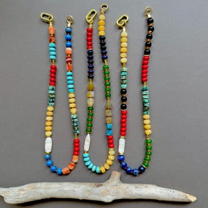 May include: Five necklaces featuring multi-coloured beads arranged in distinct patterns. Each strand includes a variety of opaque, translucent, and textured beads in shades of red, blue, green, yellow, and white, finished with a gold-toned metal loop.