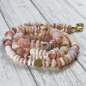 Könnte beinhalten: Eine Halskette aus rosa und weißen Edelsteinperlen mit goldfarbenen Metallakzenten. Die Perlen variieren in Größe und Form und erzeugen ein strukturiertes Aussehen. Die Halskette wird auf einer hellen Holzoberfläche präsentiert.