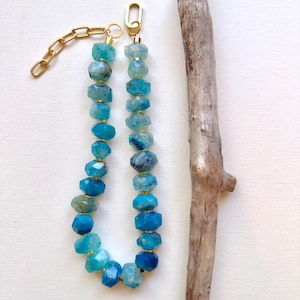 May include: A necklace featuring faceted, translucent blue gemstone beads strung together with small gold-toned spacers. The necklace includes a gold-toned metal chain extender with large oval links and a lobster claw clasp at the end.