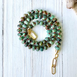 May include: A necklace with turquoise beads, speckled with brown and black, strung on a red cord. The necklace is fastened with gold-coloured clasps. The oval-shaped beads are arranged in a spiral. The necklace is displayed on a white wooden surface.