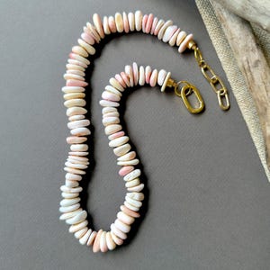 Puede incluir: Un collar compuesto por numerosas cuentas pequeñas, planas y con forma de disco en tonos blanco, crema y rosa suave. Las cuentas están ensartadas y el collar cuenta con un cierre de metal dorado con una extensión de cadena ajustable.