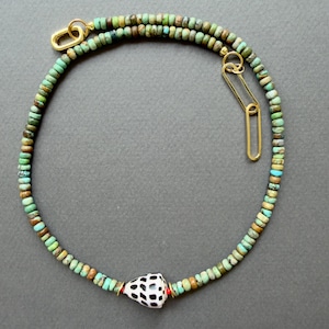 Peut inclure: Un collier composé de petites perles rondes et variées de couleur turquoise, dans des tons de vert et de brun, centré par une perle en coquillage conique blanc à taches noires. Le collier comporte des maillons dorés aux extrémités.