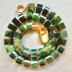 May include: A necklace composed of cylindrical, faceted dark green stone beads separated by small gold-toned metal spacers. The piece features a gold-toned circular clasp at one end and an elongated oval lobster claw clasp at the other end.