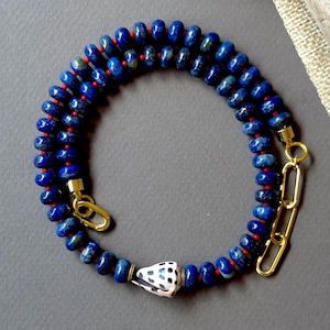 Peut inclure: Un collier composé d'une rangée de perles rondes en lapis-lazuli bleu profond, séparées par de minuscules perles rouges. Le bijou présente une breloque centrale en coquillage blanc à motifs noirs et des maillons dorés avec un fermoir.