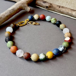 May include: A beaded bracelet featuring a variety of colorful, round gemstones. The bracelet has a gold-colored clasp and chain. The gemstones are in shades of red, blue, green, yellow, and brown.