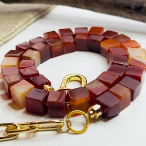 May include: A necklace with cube-shaped beads in red, orange, and amber hues. The necklace has a gold-coloured clasp and chain, contrasting with the earthy tones of the beads. The beads are approximately 1 cm in size.