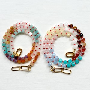 May include: Two necklaces featuring multi-colored gemstone beads. The left necklace has square-cut beads in shades of blue, orange, and purple. The right necklace has round, faceted beads in white, yellow, pink, blue, and brown, both with gold-tone links.