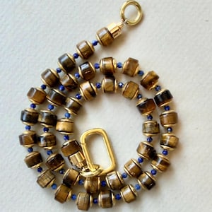 May include: A necklace featuring cylindrical brown tiger eye beads alternating with small, round, deep blue beads. The piece includes gold-toned metal spacers between the beads and is finished with a gold-toned oval carabiner-style clasp.