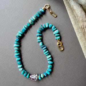 Peut inclure: Un collier composé de pépites de pierre couleur turquoise irrégulières enfilées avec de petites perles orange. Une perle en coquillage blanc à taches noires est placée vers le bas. Le collier comporte des maillons et un fermoir dorés.