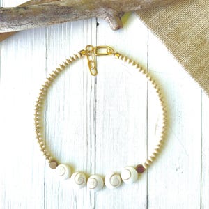 May include: A cream-colored necklace featuring a series of small, round wooden beads and larger, white shell beads. The necklace is secured with a gold-colored clasp and has a natural, beachy aesthetic.