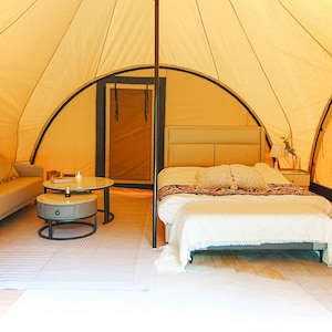 May include: A white canvas tent with a bed, couch, and side tables. The tent has a wooden frame and a large window. The bed has a white comforter and pillows. The couch is a light brown color and has a few pillows. The side tables are made of wood and have a lamp and a vase on them.