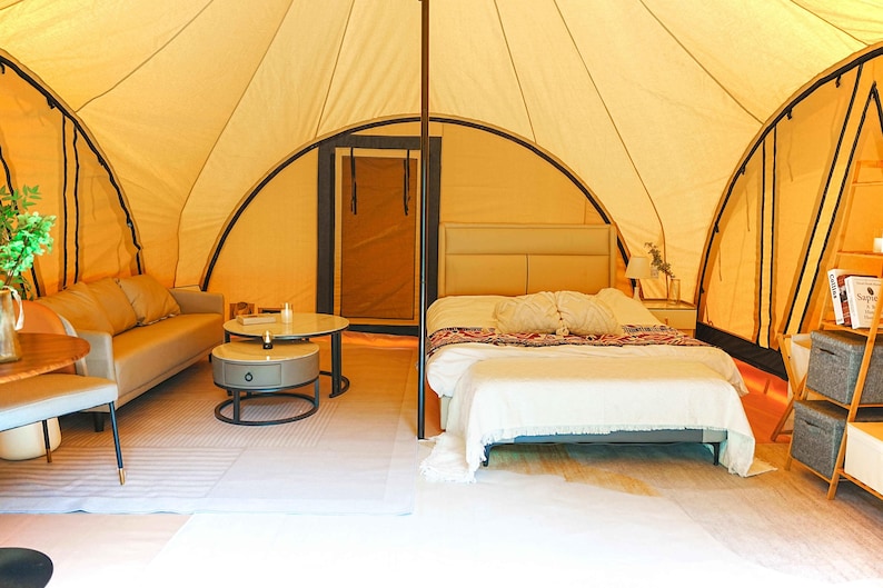 May include: A white canvas tent with a bed, couch, and side tables. The tent has a wooden frame and a large window. The bed has a white comforter and pillows. The couch is a light brown color and has a few pillows. The side tables are made of wood and have a lamp and a vase on them.
