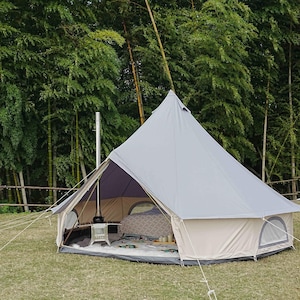 May include: A light gray bell tent pitched on a grassy area, with a beige base and a dark purple interior. The tent has a small window and is secured with ropes. A metal chimney extends from the tent's interior. The background features lush green trees.