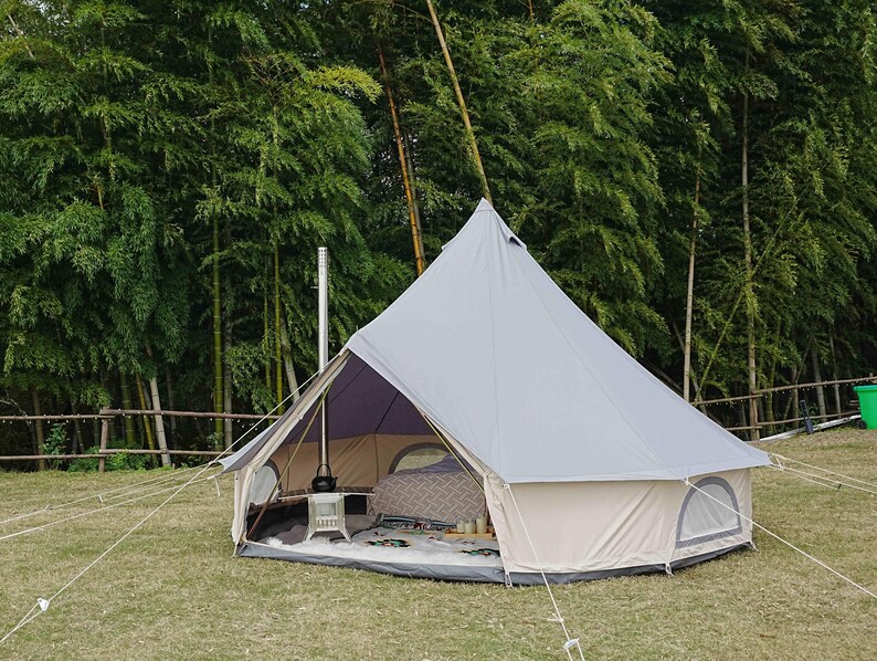 May include: A light gray bell tent pitched on a grassy area, with a beige base and a dark purple interior. The tent has a small window and is secured with ropes. A metal chimney extends from the tent's interior. The background features lush green trees.
