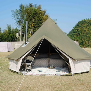 May include: A large olive green canvas tent with a conical roof, set up outdoors on a grassy area. The tent has a white base and is open, revealing a bed with patterned bedding. A small table and a rug are inside.