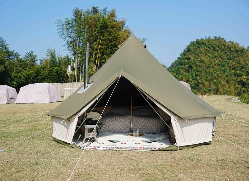 May include: A large olive green canvas tent with a conical roof, set up outdoors on a grassy area. The tent has a white base and is open, revealing a bed with patterned bedding. A small table and a rug are inside.