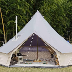 May include: A light gray bell tent pitched outdoors, featuring a central chimney and open front. Inside, a bed is made up with patterned bedding. A small stove and decorative items are visible on the floor. The tent is set up on grass with a backdrop of trees.
