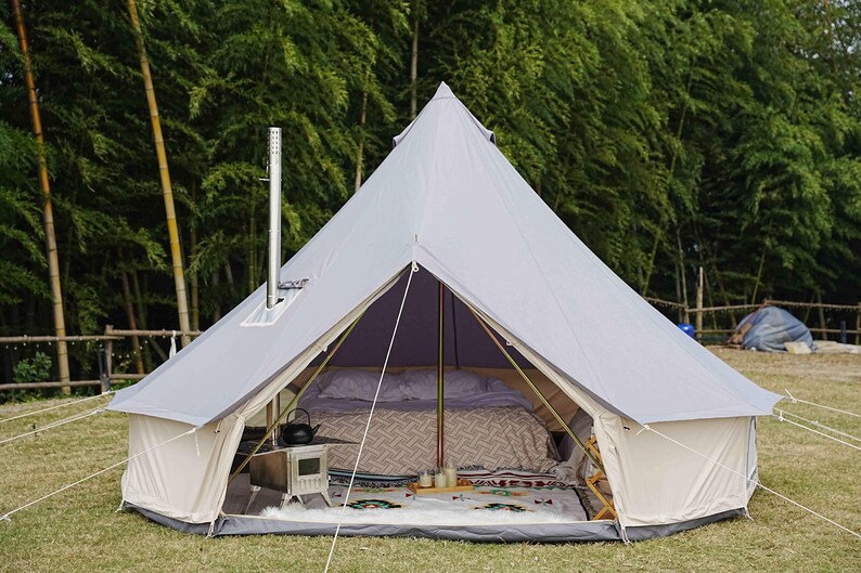 May include: A light gray bell tent pitched outdoors, featuring a central chimney and open front. Inside, a bed is made up with patterned bedding. A small stove and decorative items are visible on the floor. The tent is set up on grass with a backdrop of trees.