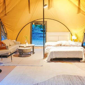 May include: A white bed with a white blanket and pillows is in a yellow tent with a wooden floor. There is a small table and chairs in the foreground.