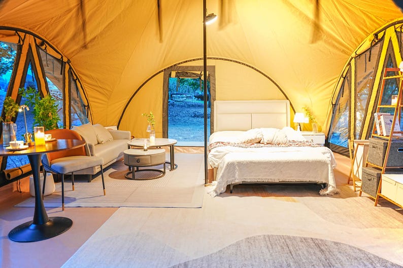 May include: A white bed with a white blanket and pillows is in a yellow tent with a wooden floor. There is a small table and chairs in the foreground.