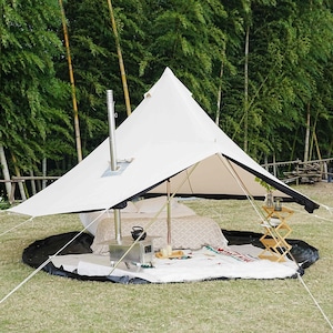 May include: A white canvas tent with a black trim, set up outdoors on a grassy area. The tent has a central pole and a chimney. Inside, there's a bed, a small stove, and a wooden side table. The background features bamboo trees.