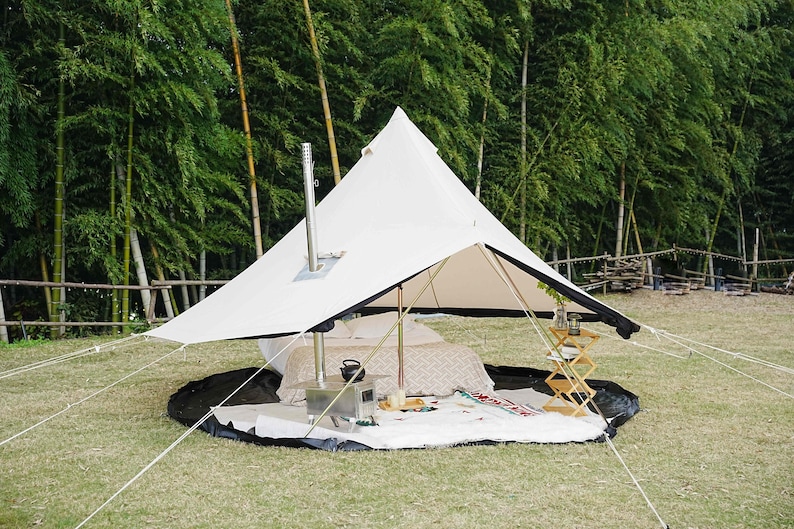 May include: A white canvas tent with a black trim, set up outdoors on a grassy area. The tent has a central pole and a chimney. Inside, there's a bed, a small stove, and a wooden side table. The background features bamboo trees.