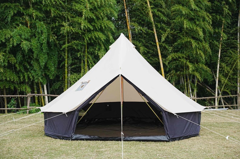 May include: A large, white canvas tent with a dark blue base and trim, set up on a grassy area. The tent has a central pole and is open at the front, revealing a dark interior. Bamboo trees are in the background.