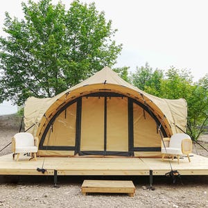 May include: A large, tan canvas tent with a wooden platform and two white chairs. The tent has a large, arched entrance and is surrounded by trees.