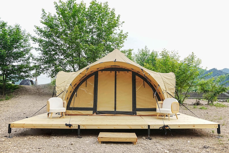 May include: A large, tan canvas tent with a wooden platform and two white chairs. The tent has a large, arched entrance and is surrounded by trees.