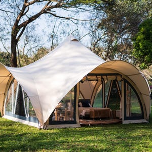 May include: A white canvas tent with a large, arched entrance and multiple windows. The tent is set up on a grassy lawn with trees in the background.