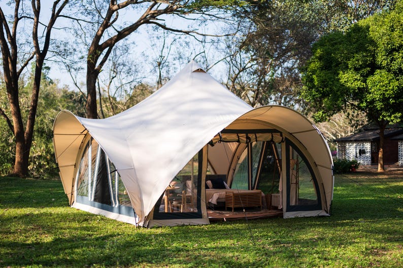 May include: A white canvas tent with a large, arched entrance and multiple windows. The tent is set up on a grassy lawn with trees in the background.