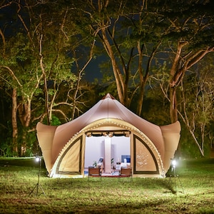 May include: A large, beige tent with a rounded roof and a large open front. The tent is lit up with string lights and has two chairs inside. The tent is set up in a grassy area with trees in the background.