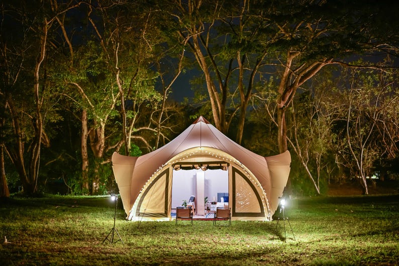 May include: A large, beige tent with a rounded roof and a large open front. The tent is lit up with string lights and has two chairs inside. The tent is set up in a grassy area with trees in the background.