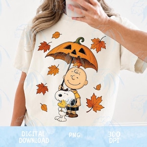 May include: A cream-colored t-shirt featuring a graphic of Charlie Brown and Snoopy standing together under a large orange pumpkin-shaped umbrella, surrounded by scattered orange autumn leaves. The design is centered on the front of the shirt.