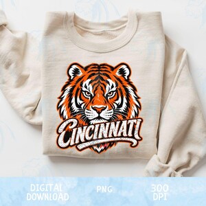 Può includere: Una felpa girocollo color crema con la grafica di un muso di tigre con motivi di pelo arancione, nero e bianco. Sotto la tigre, la parola CINCINNATI è stampata in un carattere bianco dall'aspetto usurato con un contorno nero sul tessuto.