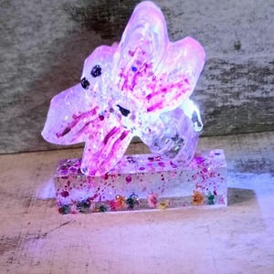 May include: A clear resin sculpture of a flower with pink and purple accents, glitter, and small colorful flowers. The flower is mounted on a rectangular base with glitter and small flowers.