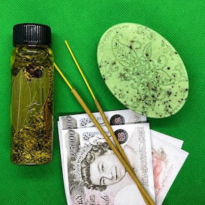 Könnte beinhalten: Eine kleine Glasflasche mit schwarzem Deckel, gefüllt mit Öl und Kräutern, eine grüne ovale Seife mit floralem Design, zwei Räucherstäbchen und Geldscheine auf grünem Hintergrund.