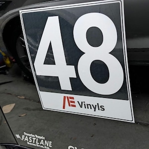 Puede incluir: Una pegatina negra y blanca con el número 48 en un coche negro. La pegatina tiene el texto "NE Vinyls" en rojo y blanco.