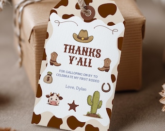 May include: A rectangular paper gift tag with a brown and white cow print border. The tag features a brown cowboy hat, boots, a money bag, a horseshoe, a cow face, a star, and a cactus. Text reads: THANKS Y'ALL FOR GALLOPING ON BY TO CELEBRATE MY FIRST RODEO Love, Dylan.